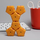 The Speks Geode: Solar Flare is a bright yellow fidget toy made of interconnected hexagonal pieces with red connectors. It is designed for tactile stimulation and creativity, allowing users to build various shapes. The toy is positioned on a clean workspace with a keyboard and a pair of scissors in a red cup in the background.