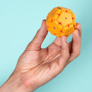 A person holds the Speks Geode: Solar Flare, a vibrant yellow and red spherical puzzle toy, featuring interlocking hexagonal and pentagonal shapes, against a pastel blue background.