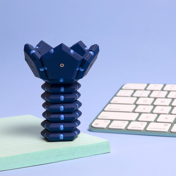 The Speks Geode: Space Cadet is a unique fidget toy featuring a stackable design in dark blue and light blue colors. It consists of a hexagonal top and a segmented base that can be manipulated for stress relief and creative play. The background includes a light blue setting with a notepad and an off-white keyboard.