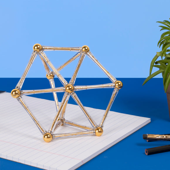 A geometric structure made of clear rods and golden spheres, placed on a lined notebook. This decorative item by SPEKS combines creativity with engineering. Ideal for stress relief and enhancing focus.