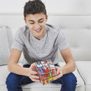 A young boy sitting on a light-colored couch, smiling as he holds the Perplexus Rubik's Fusion 3x3 mechanical puzzle. The puzzle is colorful, showcasing a mix of different pieces within a transparent cube. The boy is wearing a gray t-shirt and denim jeans, demonstrating engagement with the challenging puzzle.