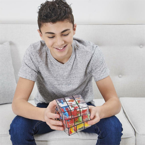 A young boy sitting on a light-colored couch, smiling as he holds the Perplexus Rubik's Fusion 3x3 mechanical puzzle. The puzzle is colorful, showcasing a mix of different pieces within a transparent cube. The boy is wearing a gray t-shirt and denim jeans, demonstrating engagement with the challenging puzzle.