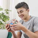 A boy with short dark hair smiles as he holds and examines the Perplexus Rubik's Fusion 3x3 mechanical puzzle. The puzzle features vibrant colors and geometric designs, captivating his attention. In the background, there are green plants and a softly lit room, creating a warm atmosphere.