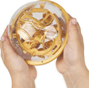 A person holds the Harry Potter Perplexus Golden Snitch, a mechanical puzzle featuring a translucent outer shell that reveals intricate inner pathways and mechanisms in a golden color. The design resembles the iconic Golden Snitch from the Harry Potter series.
