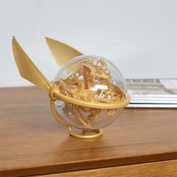A detailed view of the Harry Potter Perplexus Golden Snitch by Spin Master. The mechanical puzzle features a transparent globe with intricate pathways and a gold-colored outer structure resembling the iconic snitch from the wizarding world, complete with distinct wings. It is displayed on a wooden surface.