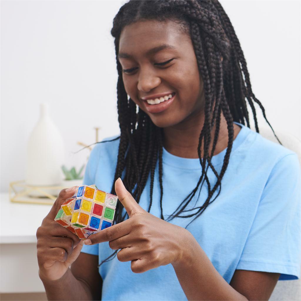 A young girl with braided hair smiles as she plays with the Rubik's 3x3 Crystal Cube from Spin Master. The cube features vibrant, colorful tiles reflecting light, making it visually captivating. She is focused on solving the mechanical puzzle, demonstrating both concentration and enjoyment.