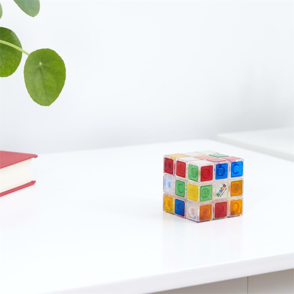 A colorful Rubik's 3x3 Crystal Cube by Spin Master displayed on a white table. The cube features transparent colored squares that allow for visibility of the internal mechanism. A plant and a red book are partially visible in the background, creating a soothing workspace setting. The cube is designed to challenge and entertain users with its mechanical puzzle format.