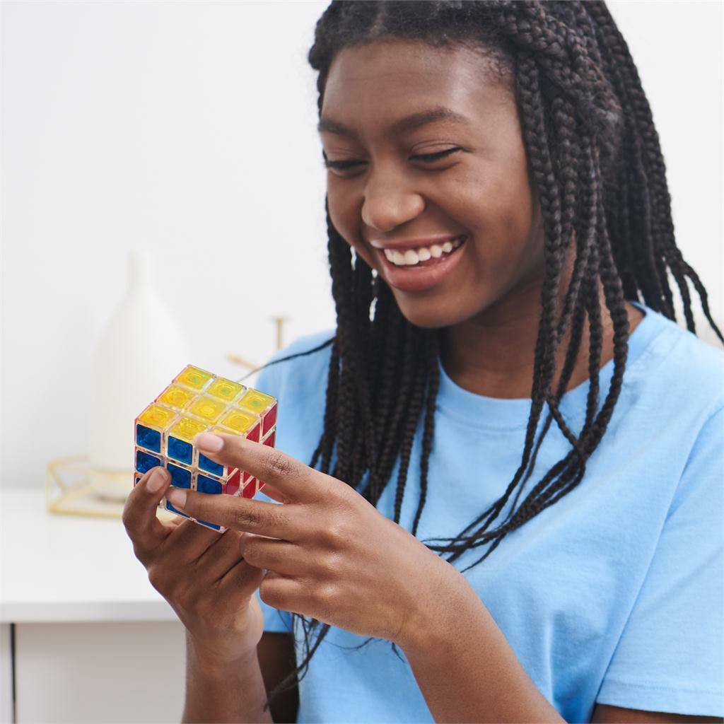 A young girl with braided hair smiles joyfully while holding a Rubik's 3x3 Crystal Cube. She is focused on the colorful mechanical puzzle in her hands, showcasing its vibrant yellow, red, blue, and clear plastic squares. The background features a modern, minimalist decor.