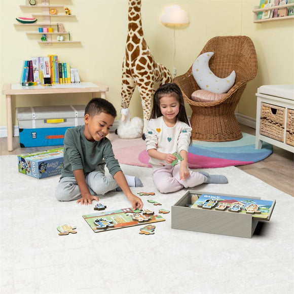 Two children playing with the Bluey 4 Wood Puzzles jigsaw set on a soft rug. They are joyfully assembling puzzle pieces featuring colorful characters from the Bluey series. A box containing the puzzles is visible in the background, along with cheerful decor in a bright playroom.