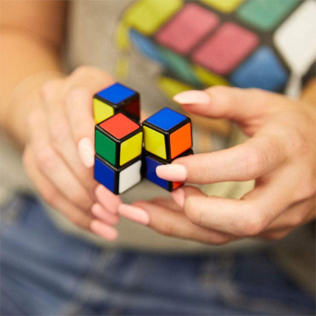 A person is holding the Rubik's 3x1 Edge mechanical puzzle in their hands, featuring colorful squares on three smaller cubes that connect together. The background shows a blurred image of a casual setting, highlighting the puzzle's vibrant colors against the wearer's t-shirt.