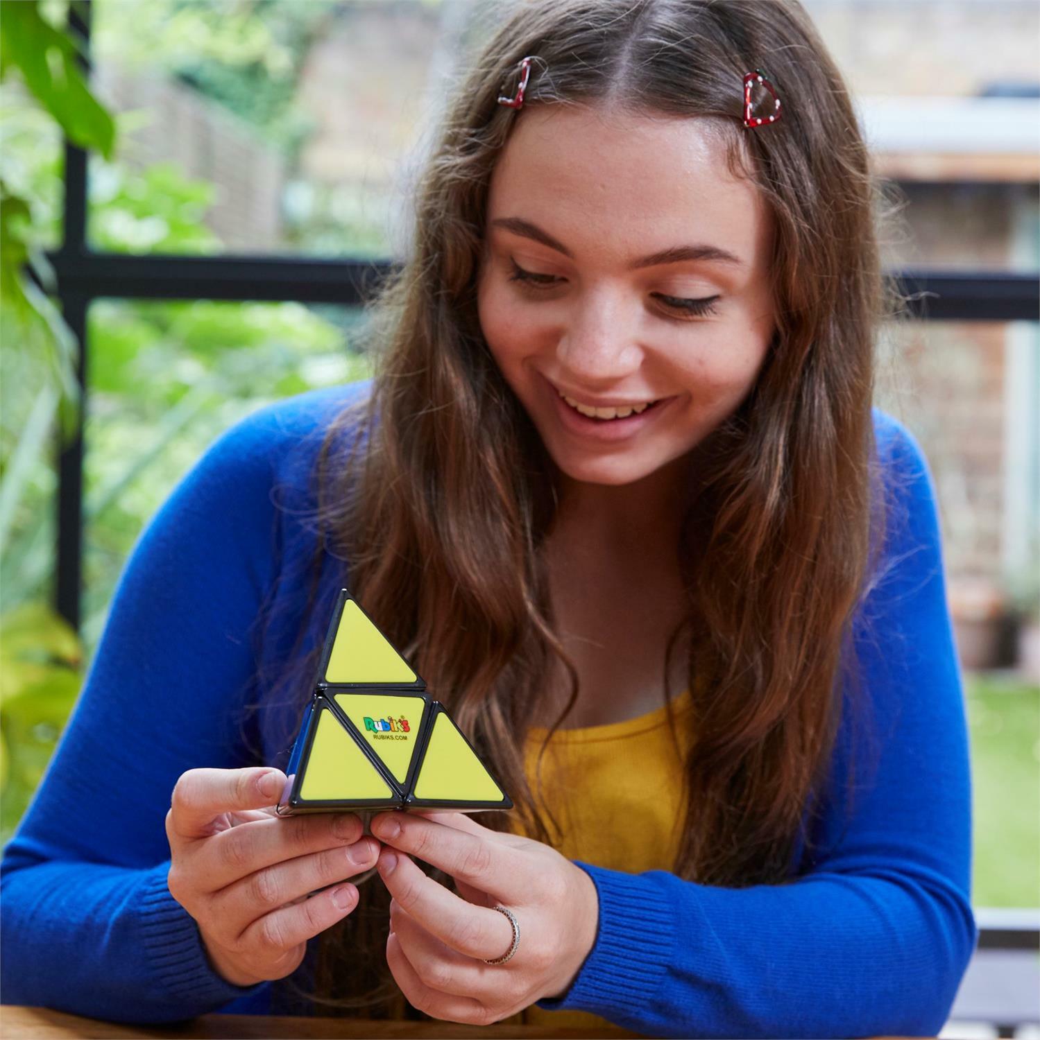 A young woman with long, wavy hair and wearing a blue cardigan smiles while manipulating a Rubik's Pyramid puzzle made by Spin Master. The puzzle has a triangular shape with bright yellow pieces. She is sitting at a table in a bright room with lush greenery in the background.