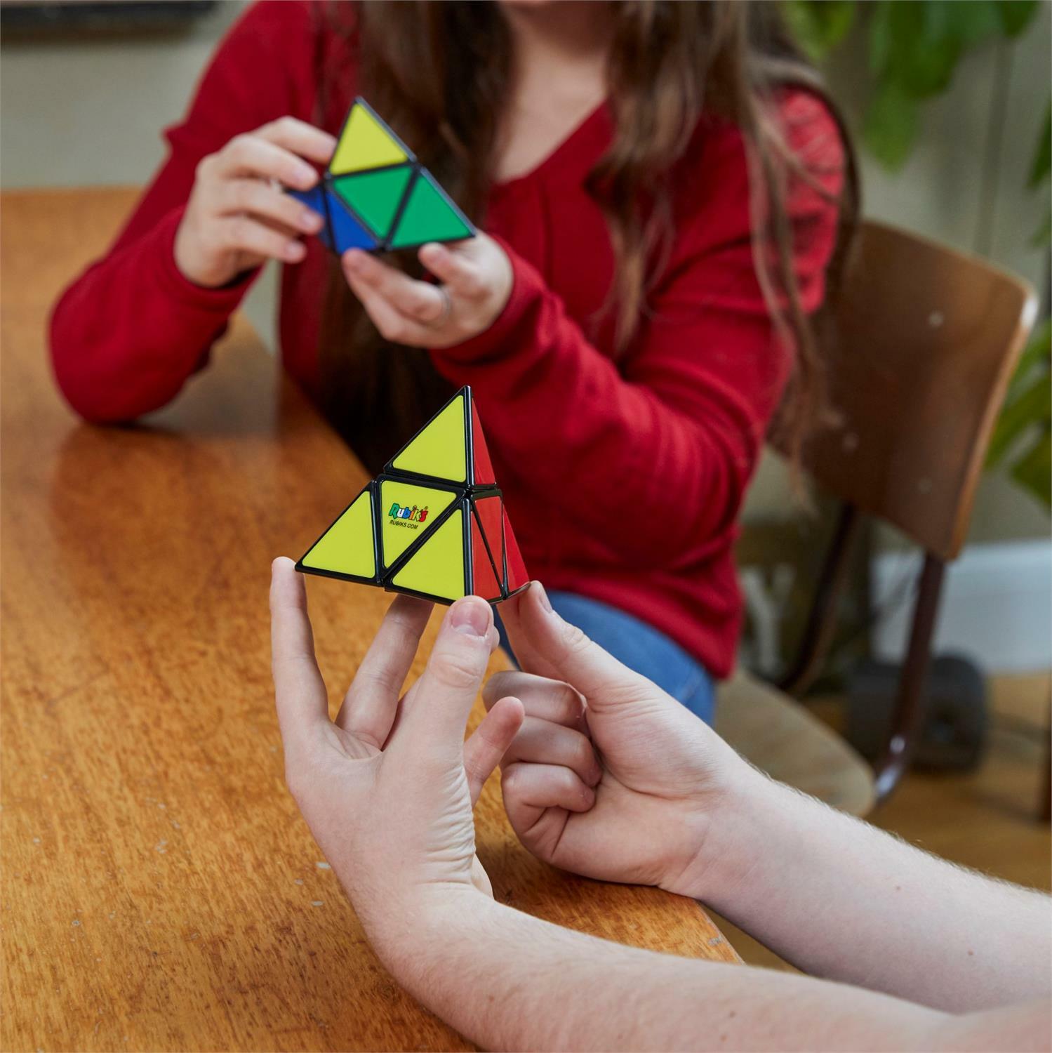 Two people engaged in solving the Rubik's Pyramid puzzle from Spin Master, showcasing a colorful triangular design with yellow, green, blue, and red segments on a wooden table.