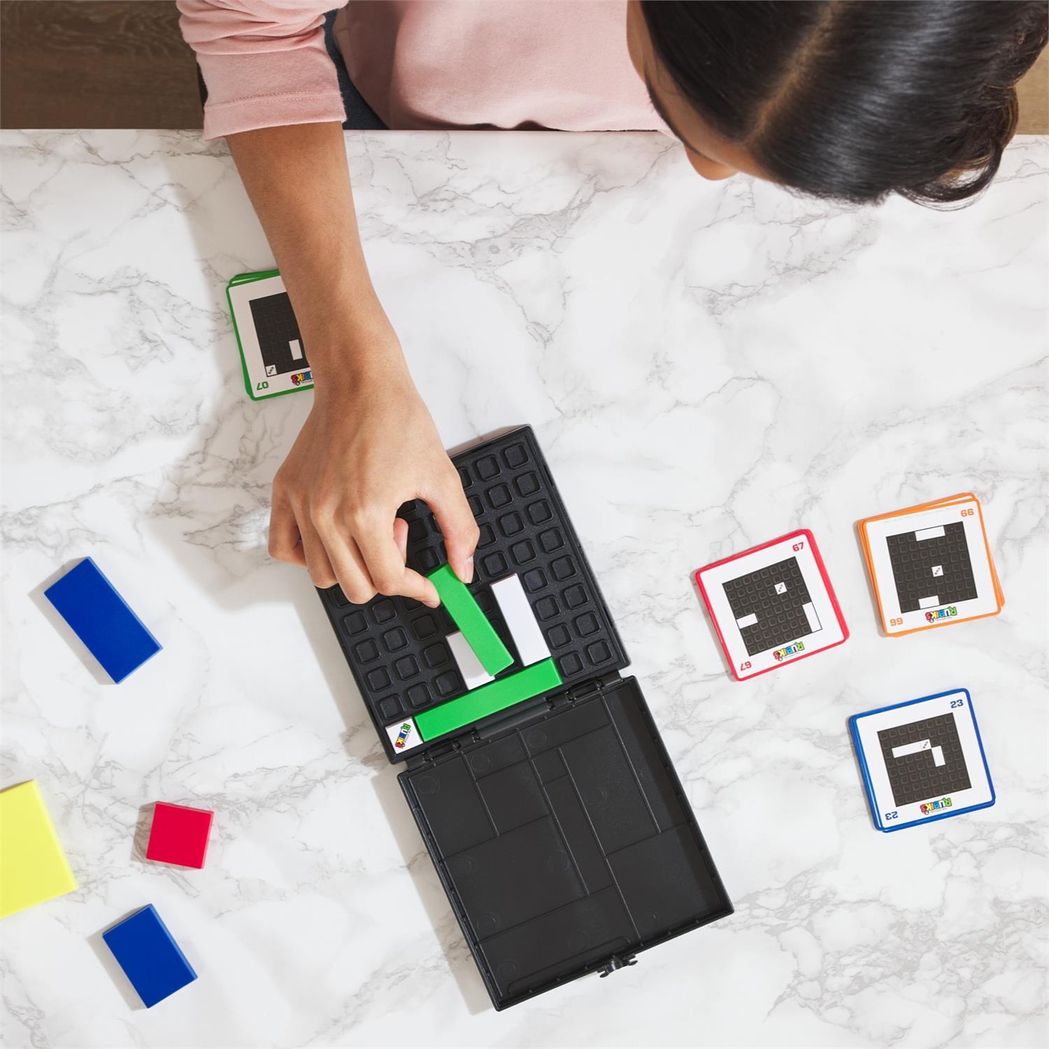 A person solving the Rubik's Cube Gridlock Game by Spin Master, featuring a black game board with a green piece being maneuvered into a slot. Nearby are colorful blocks and challenge cards in red, blue, and green.