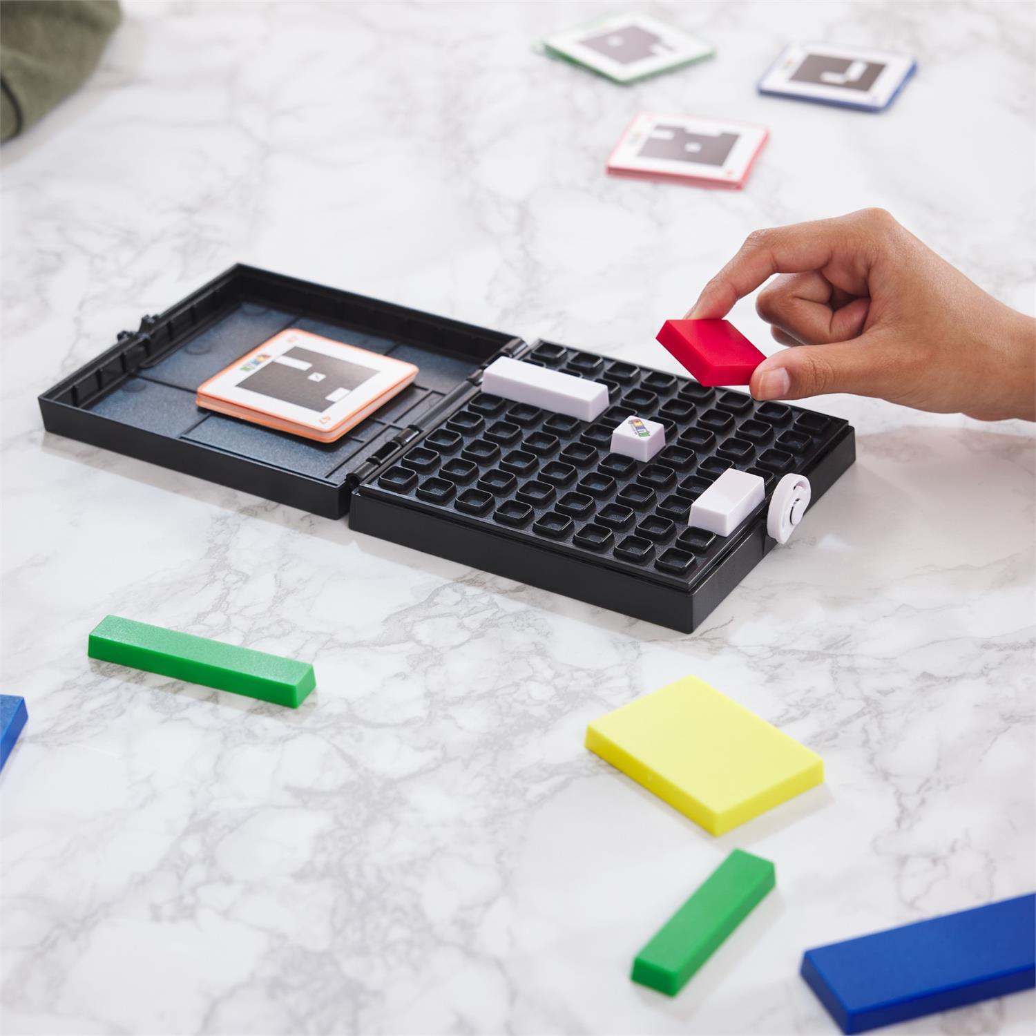 A person is placing a red tile onto a black grid of the Rubik's Cube Gridlock Game by Spin Master. The game includes colorful tiles in various shapes and a set of task cards to guide players. The grid is open on a marble-patterned surface, showcasing other colorful tiles in green, yellow, and blue around it.