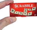 A hand holding a compact version of Scrabble game titled 'World's Smallest Scrabble' by Super Impulse. The red game box features letters on tiles spelling 'PLAYS' and includes scoring details from the original game.