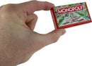 A person holding the World's Smallest Monopoly game, showing the tiny red box featuring the game's logo and Monopoly board artwork. The game is compact and fits in the palm of the hand.