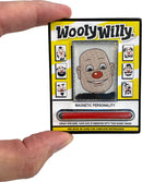 A person holding the World's Smallest WoolyWilly toy by Super Impulse. The toy features a smiling character with a red nose and various face designs. It includes a magnetic wand to create whiskers, hair, and eyebrows using iron filings, all within a colorful package.