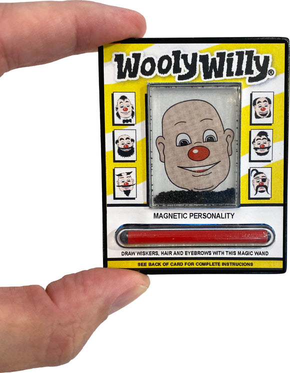 A person holding the World's Smallest WoolyWilly toy by Super Impulse. The toy features a smiling character with a red nose and various face designs. It includes a magnetic wand to create whiskers, hair, and eyebrows using iron filings, all within a colorful package.