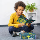A child sitting on the floor in a bright yellow shirt, reading an instruction booklet for the Gecko Run: Marble Run kit. The child is engaged and focused, with the kit box open beside them. The background features a green plant in a pot, contrasting with the neutral space.