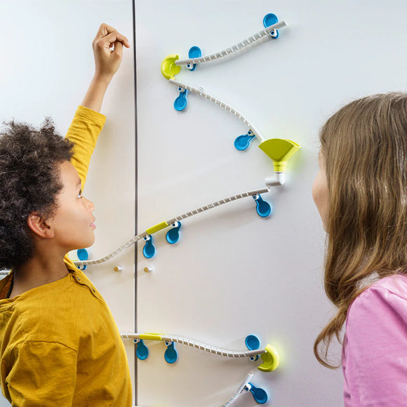 Two children play with the Gecko Run: Marble Run DIY kit by Thames & Kosmos, engaging with colorful tracks and funnels mounted on a wall, showcasing creativity and fun.