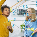 Two children engaged in a hands-on activity with the Gecko Run: Marble Run DIY kit by Thames & Kosmos. One child, wearing a yellow sweatshirt, is reaching to connect a piece, while the other, in a blue hoodie, looks up with curiosity as they build a colorful marble run together. The setup features various white and blue tubes and colorful connectors, highlighting creativity and collaboration in play.