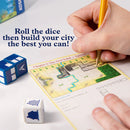 A close-up view of a hand holding a pencil poised to draw on a game sheet for 'My City Roll & Build' by Thames & Kosmos. Two colorful dice are visible, featuring icons related to the game, positioned next to the game board. The sheet shows a grid layout with natural elements like rivers and trees, indicating a city-building theme. The text 'Roll the dice then build your city the best you can!' is prominently displayed.