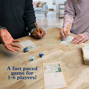 Two people playing the My City Roll & Build game at a wooden table, with a focus on game cards and dice. One player is holding a pencil while gesturing, and the other is writing on a game card. The scene is inviting and shows the interactive nature of the game, emphasizing fun and collaboration.