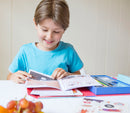 A young boy is sitting at a table, smiling while reading a comic book. He is focused on colorful pages filled with illustrations and stickers, with a blue box containing art supplies next to him. A bowl of grapes sits in the foreground, adding a bright touch to the scene.