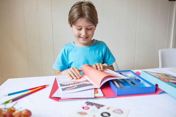 A young boy sitting at a table, engrossed in a Comic Book Kit by Tiger Tribe. He is flipping through colorful pages filled with illustrations and has various art supplies, including colored pencils and more, nearby. The scene displays creativity and fun, as the boy smiles while engaging with the kit.