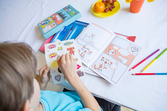 A child is engaged in creative play with the Tiger Tribe Comic Book Kit. The colorful kit includes a comic book with blank panels, various stickers featuring action words like 'POW!' and 'BANG!', and colored pencils. The workspace is lively with fruits and flowers, adding to the vibrant atmosphere of artistic expression.