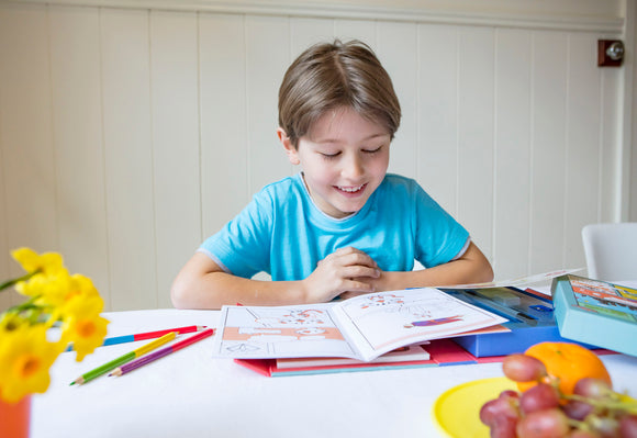 A young boy in a blue shirt smiles while reading a comic book kit from Tiger Tribe. The table is decorated with colorful drawing tools, a plate of fruit, and a vibrant flower pot, suggesting creativity and fun.