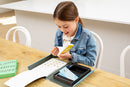 A young girl in a denim jacket joyfully exploring the Flip Book Kit by Tiger Tribe. She sits at a wooden table, holding a colorful booklet with a big smile, while the kit's components are displayed around her, showcasing creativity and fun.