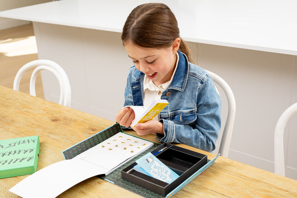 A young girl in a denim jacket joyfully exploring the Flip Book Kit by Tiger Tribe. She sits at a wooden table, holding a colorful booklet with a big smile, while the kit's components are displayed around her, showcasing creativity and fun.