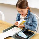 A young girl smiles as she holds a Flip Book from Tiger Tribe, showcasing her excitement while sitting at a wooden table. The kit features a colorful design and compartments for organizing materials.