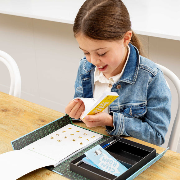 A young girl smiles as she holds a Flip Book from Tiger Tribe, showcasing her excitement while sitting at a wooden table. The kit features a colorful design and compartments for organizing materials.