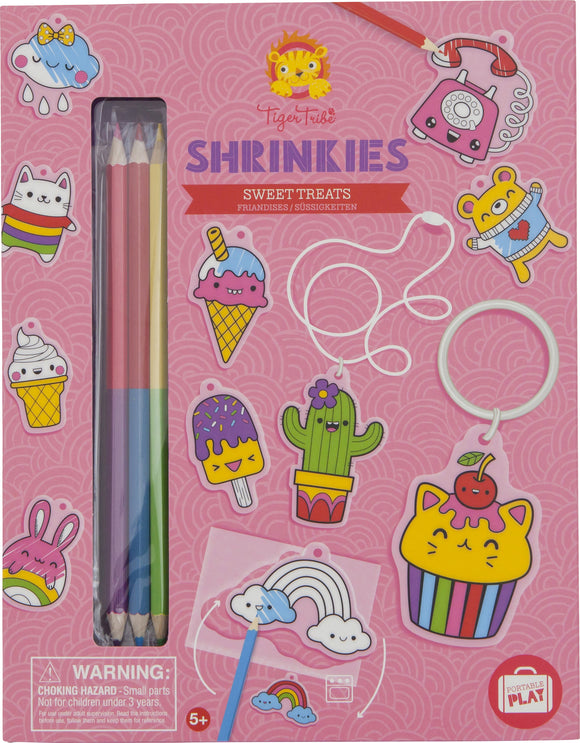 A colorful DIY kit called Sweat Treats Shrinkies by Tiger Tribe. The design features various adorable, cartoonish food characters, including ice cream cones, a cupcake, a cactus, and rainbow elements. The kit includes colored pencils for drawing and shrinking the designs, along with accessories like a keyring and strings. The vibrant pink packaging ensures an engaging crafting experience for children aged 5 and above.