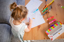 A young child with a ponytail coloring on white paper with vibrant Junior Markers from Tiger Tribe. The scene shows a wooden table covered with colored markers and some paper drawings in a bright, playful environment.