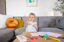 A young girl seated on a gray couch holds up a purple marker, smiling. A wooden table in front has various colorful markers scattered around, along with sheets of paper featuring drawings. Soft pillows adorn the couch, enhancing the cozy atmosphere.