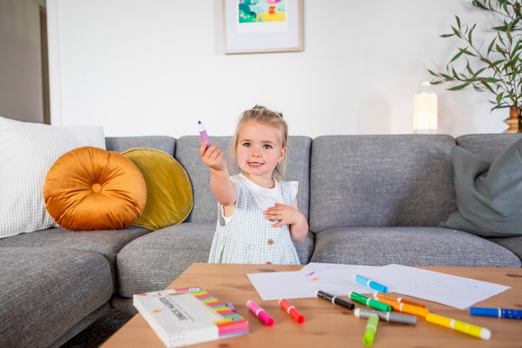A young girl seated on a gray couch holds up a purple marker, smiling. A wooden table in front has various colorful markers scattered around, along with sheets of paper featuring drawings. Soft pillows adorn the couch, enhancing the cozy atmosphere.