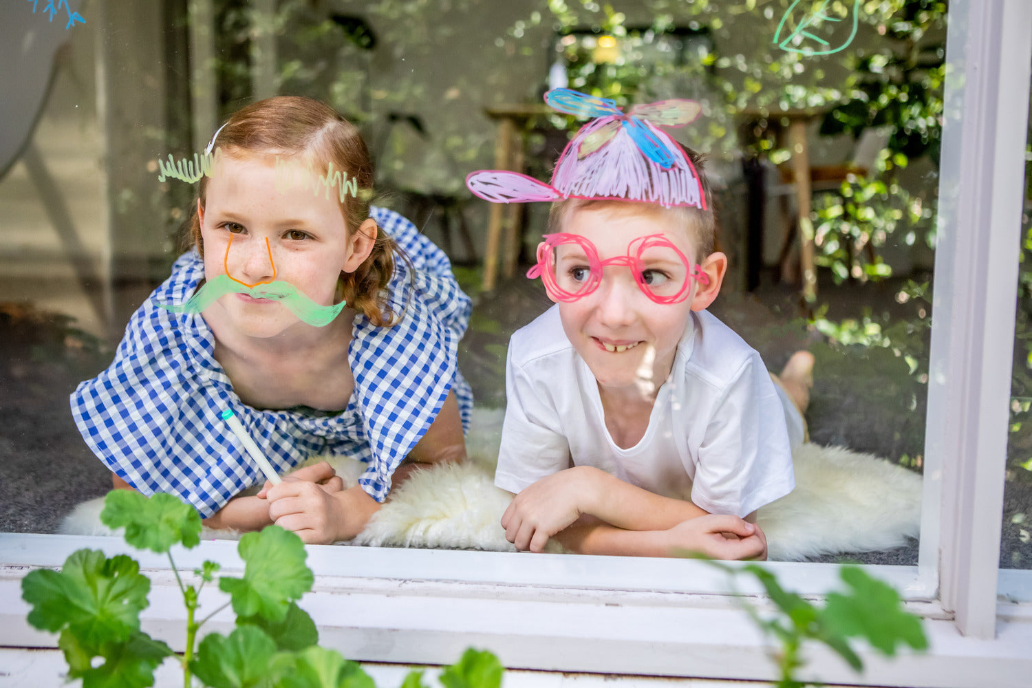 Two children playing with colorful window markers, decorating a glass window with fun designs. One girl wears a checkered dress and a playful green mustache, while the boy beside her sports oversized pink glasses and a vibrant hat drawn on the window. Their imaginative expressions and creativity are evident as they engage in this artistic activity.