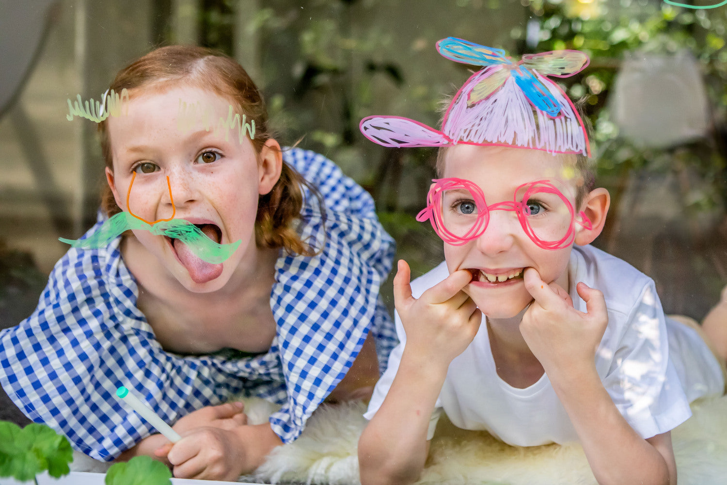 Two children playfully decorating a window using colorful window markers. One child is sticking out their tongue with a green mustache drawn on the glass, while the other is making a silly face, wearing oversized pink glasses and a colorful hair accessory. Bright and cheerful scene, showcasing creativity and fun.