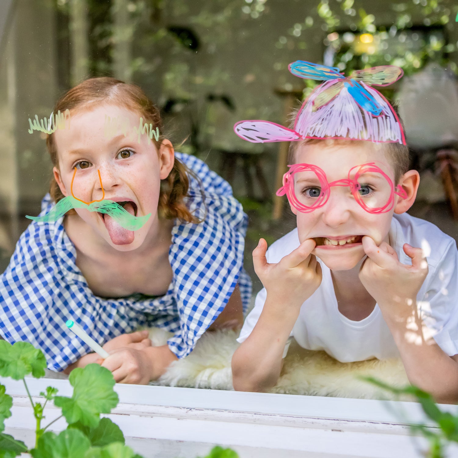 Two children having fun with colorful window markers, creating playful designs on a glass window. One child wears a green mustache and orange nose, while the other sports round pink glasses and a pink hat drawn with the markers. Lush green plants are seen in the foreground.