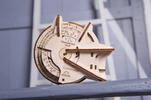 The UGears 3D Curvimeter is an intricate wooden DIY kit used for measuring curved surfaces. It features a dial with various measurement scales, including inches, centimeters, and different angles. The elegant design highlights the craftsmanship and precision that goes into making this mechanical tool, showcasing the colorful engraved details on its surface.