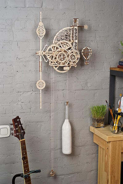 The UGears 3D Aero Clock w/Pendulum is an intricate DIY kit that features a wooden mechanical clock design with gears, pendulum, and decorative elements like a hot air balloon. It hangs elegantly on a brick wall, complementing a rustic interior with a guitar and potted plants nearby.