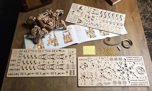 The Ugears 3D Hexapod Explorer DIY kit displayed on a wooden table. The kit includes intricately designed wooden parts, an instruction booklet, and some rubber bands. The assembled hexapod is visible, showcasing its mechanical structure, ideal for engaging creativity and building skills.