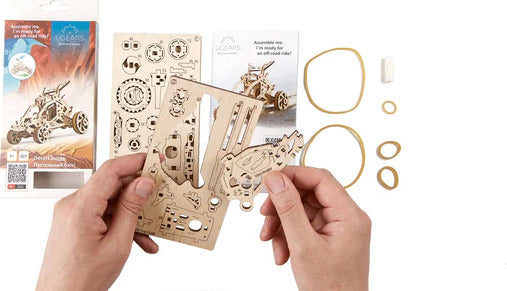 Hands holding a wooden model kit for the Ugears 3D Mini Desert Buggy. The kit includes intricate pre-cut wooden pieces, rubber bands, and assembly instructions. Ideal for DIY enthusiasts, this engaging mechanical model comes from UGears 3D.