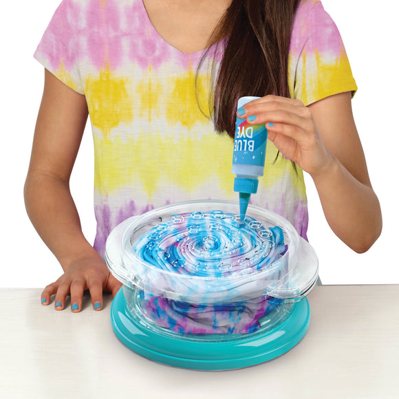 A young girl in a colorful tie-dye shirt uses a blue dye bottle over a spinning tie-dye design kit. The kit features layered fabric and a clear top, allowing the dye to blend into various swirls and patterns.