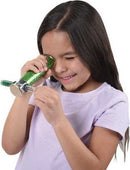 A young girl playfully looks through a green Glitter Wand Kaleidoscope, smiling as she enjoys the colorful patterns created inside. The kaleidoscope features sparkling elements that capture her attention, showcasing a fun DIY experience.