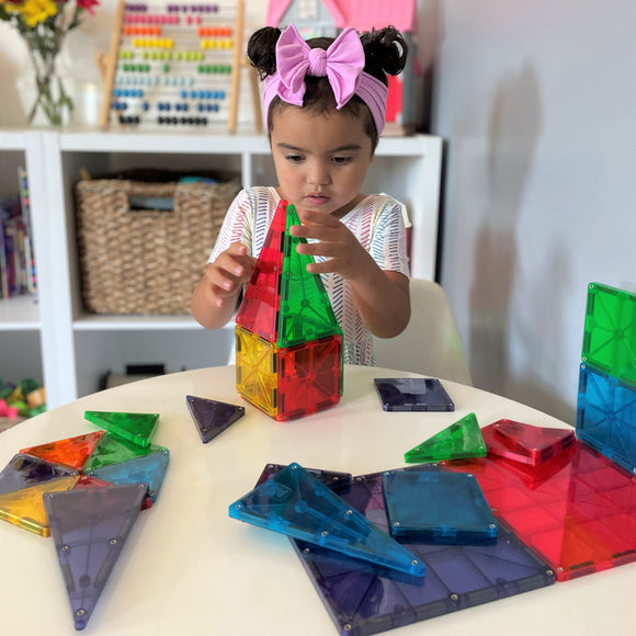 A young child playing with a 32-piece set of Magna Tiles Clear Colors. The child is focused on assembling a colorful tower using transparent geometric shapes, including red, green, yellow, and blue tiles. Various other tiles are scattered around on the table, showcasing the set's versatility. The background features organized shelves with educational toys.