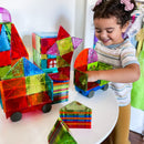 A child is joyfully playing with the Magna Tiles Metropolis 110 piece set, building colorful structures and vehicles on a white table. The tiles are translucent in various vibrant colors, including red, green, blue, and purple. The child's hand reaches into one of the bright green vehicles as they create imaginative designs, showcasing the engaging and educational nature of the product.